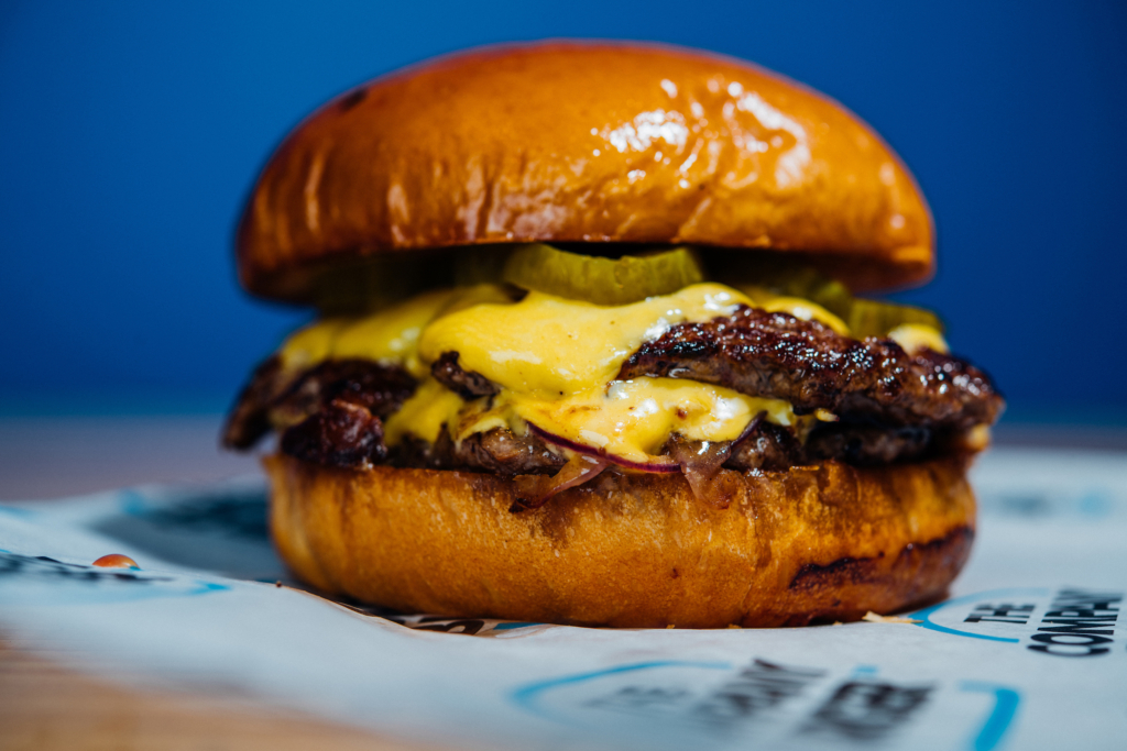 The Company Burger itself: two thin, griddled patties stacked with American cheese, bread-and-butter pickle, and red onion on a house-baked bun